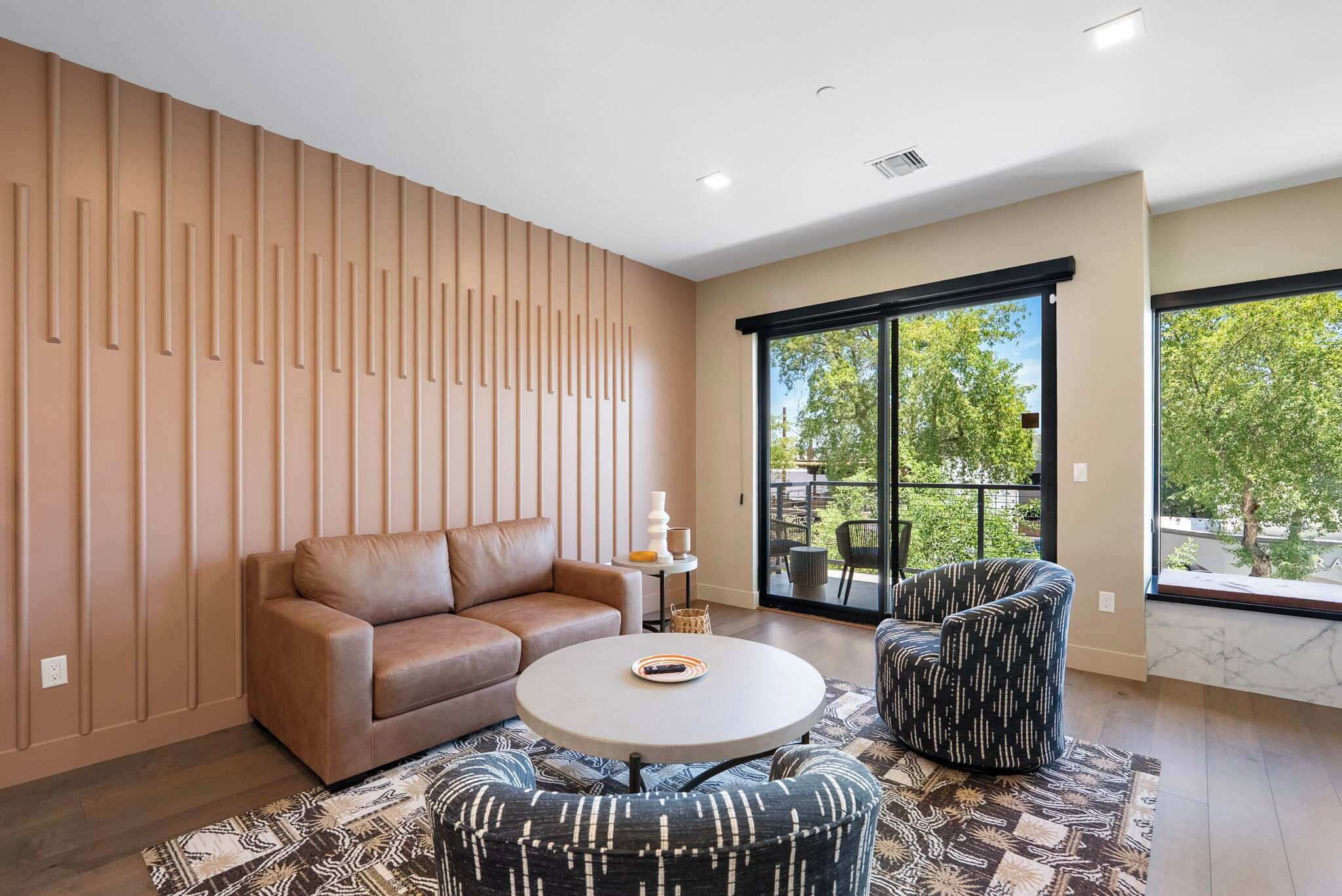 Ein modernes Wohnzimmer mit Sofa, Sessel und Couchtisch, großen Fenstern mit Blick auf Grünflächen und einem gemusterten Teppich.