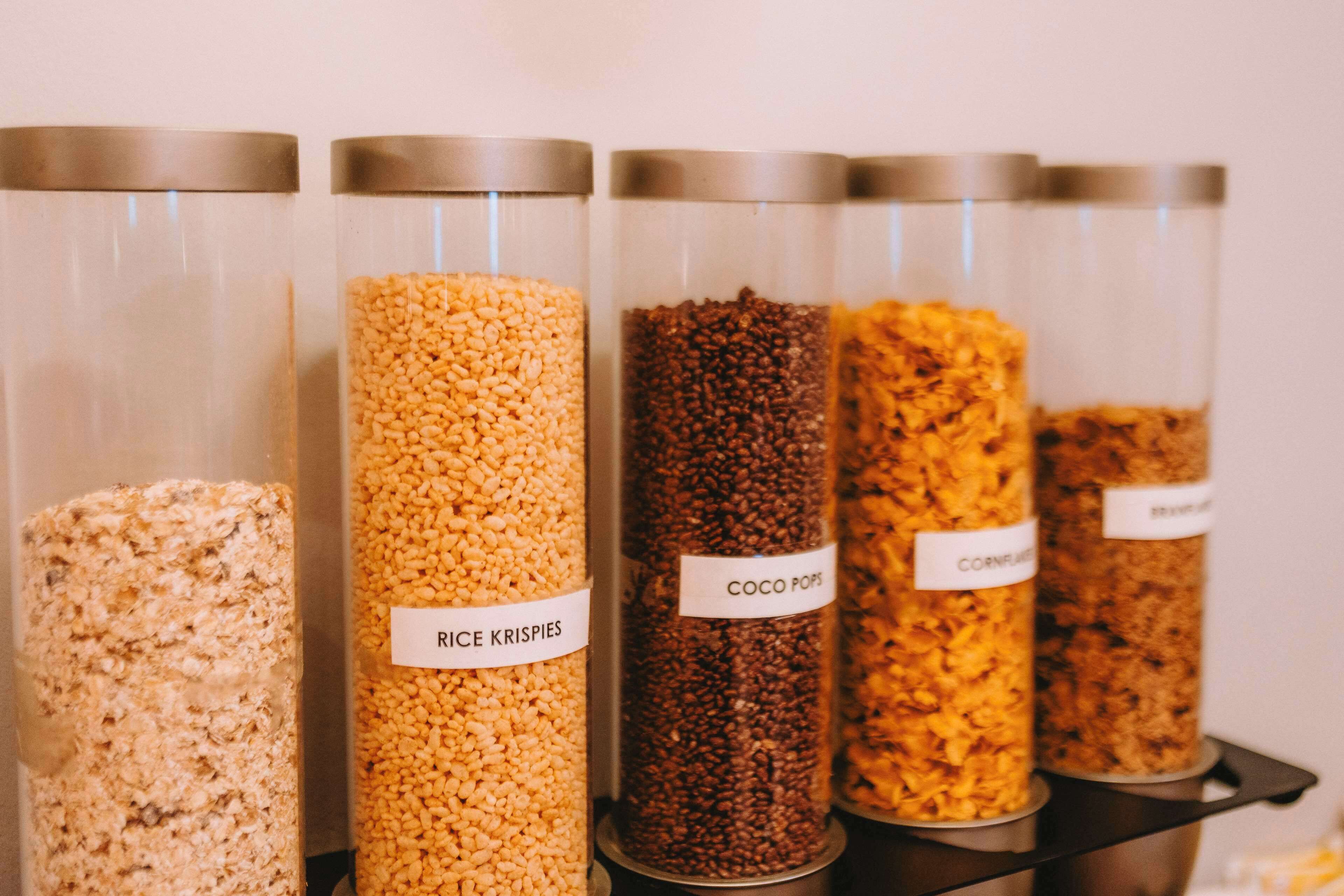 Cuatro frascos de vidrio con tapas metálicas, cada uno etiquetado con un tipo diferente de cereal: Rice Krispies, Coco Pops, Corn Flakes y otro cereal.