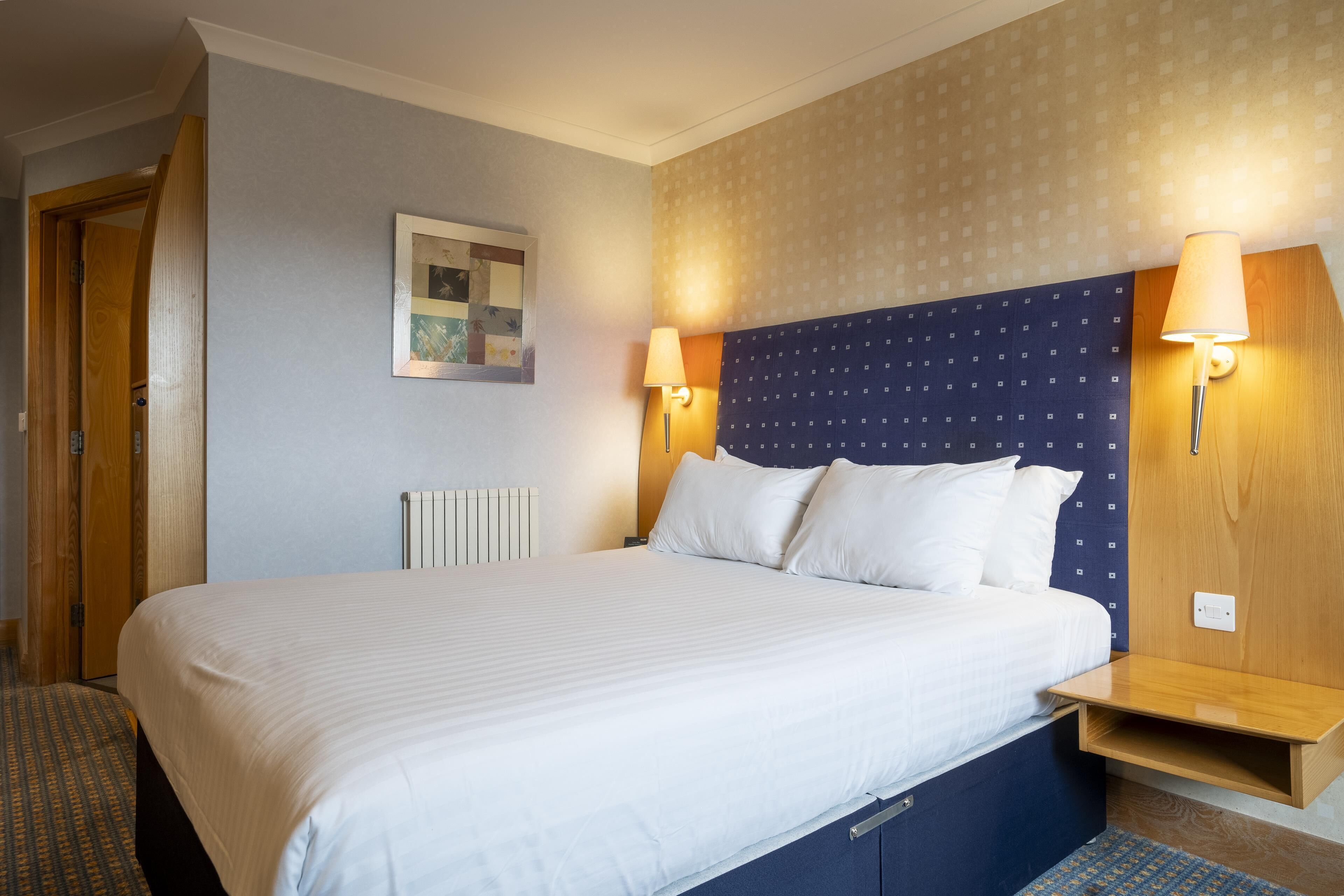 Une chambre d’hôtel avec un grand lit, deux tables de chevet avec des lampes, une tête de lit en bois et une œuvre d’art accrochée au mur.