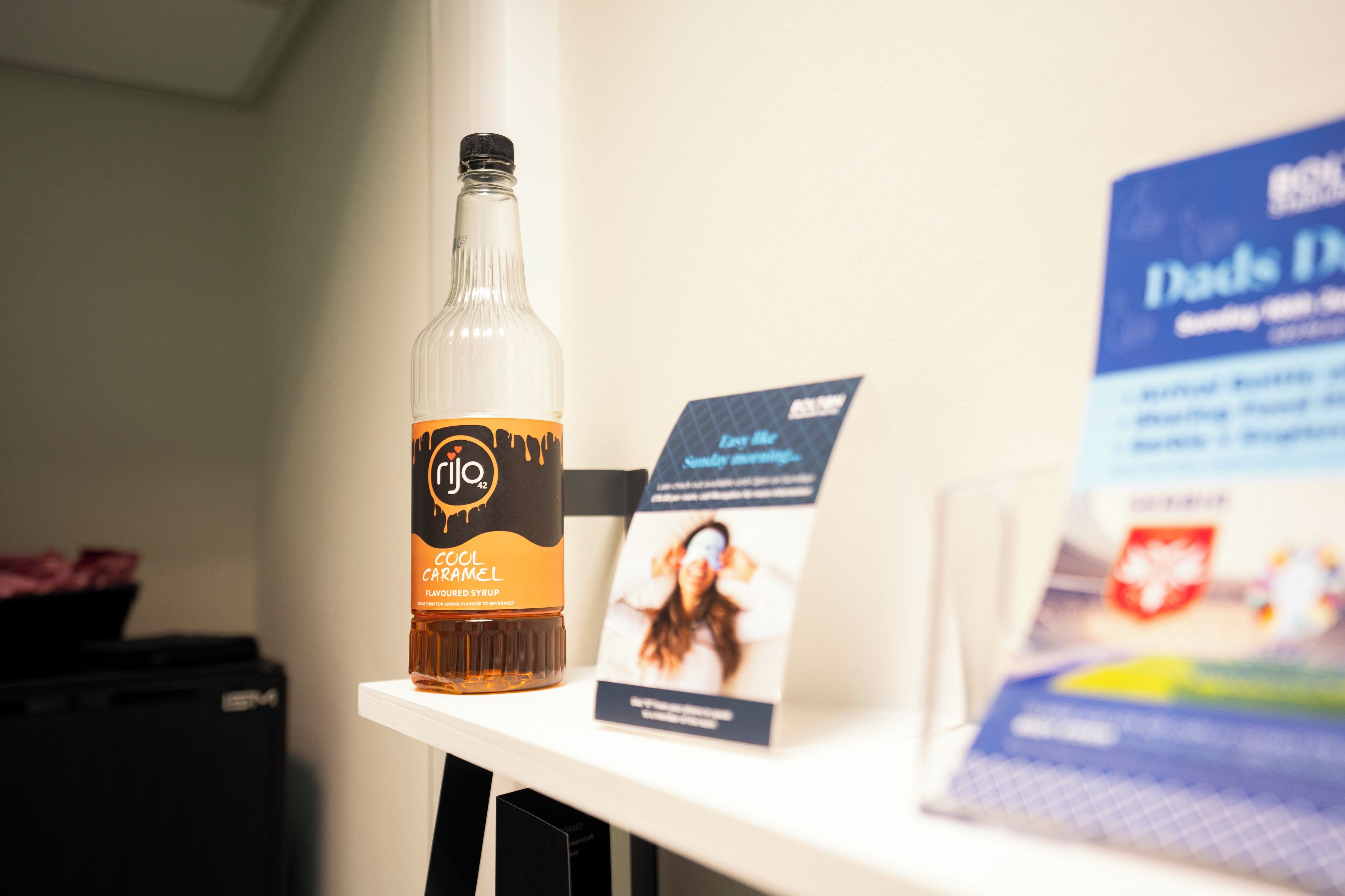 A bottle of caramel flavored syrup on a white shelf with a sign next to it.