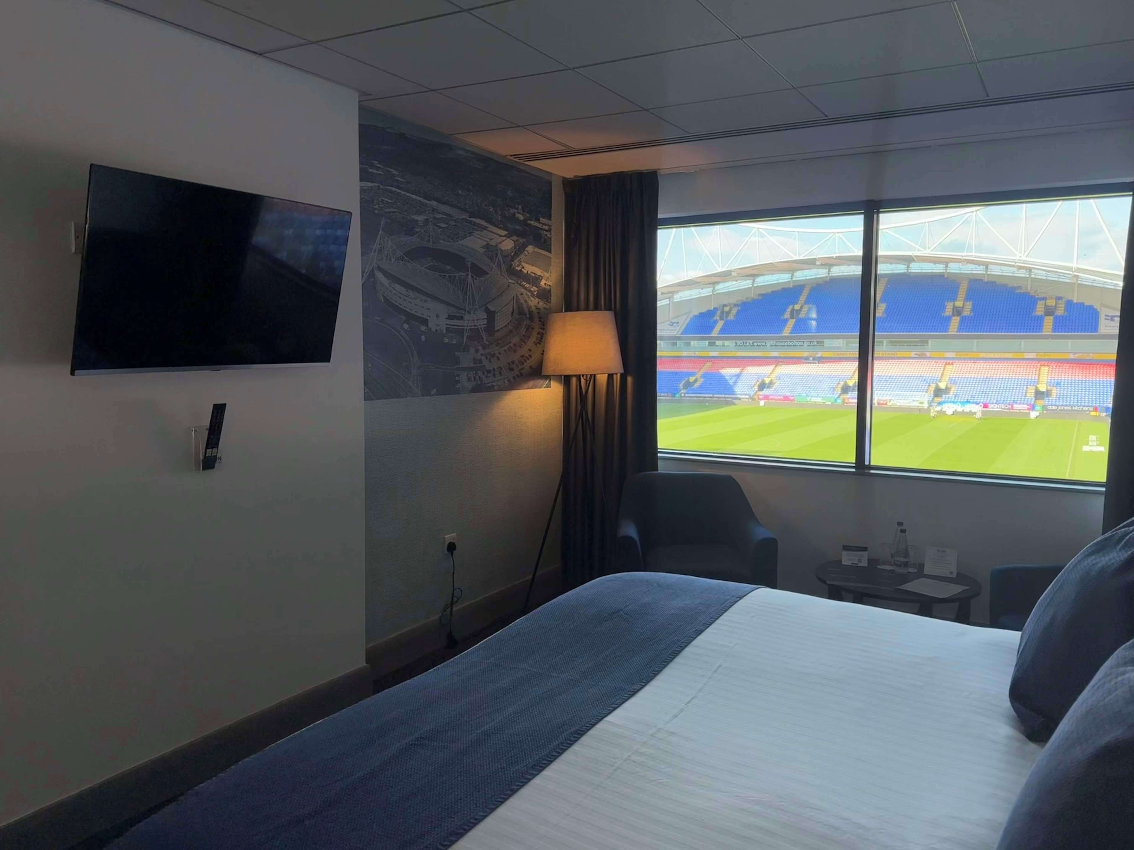 A hotel room with a bed, a TV mounted on the wall, a lamp, a window with a view of a stadium.