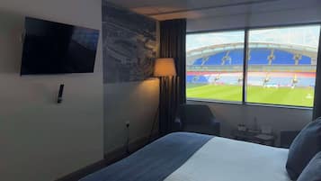 A hotel room with a bed, a TV mounted on the wall, a lamp, a window with a view of a stadium.