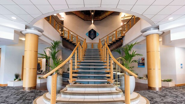 A grand staircase with wooden railings and a central column, surrounded by potted plants and a polished floor. - The Bolton Stadium Hotel, a member of Radisson Individuals (Bolton)