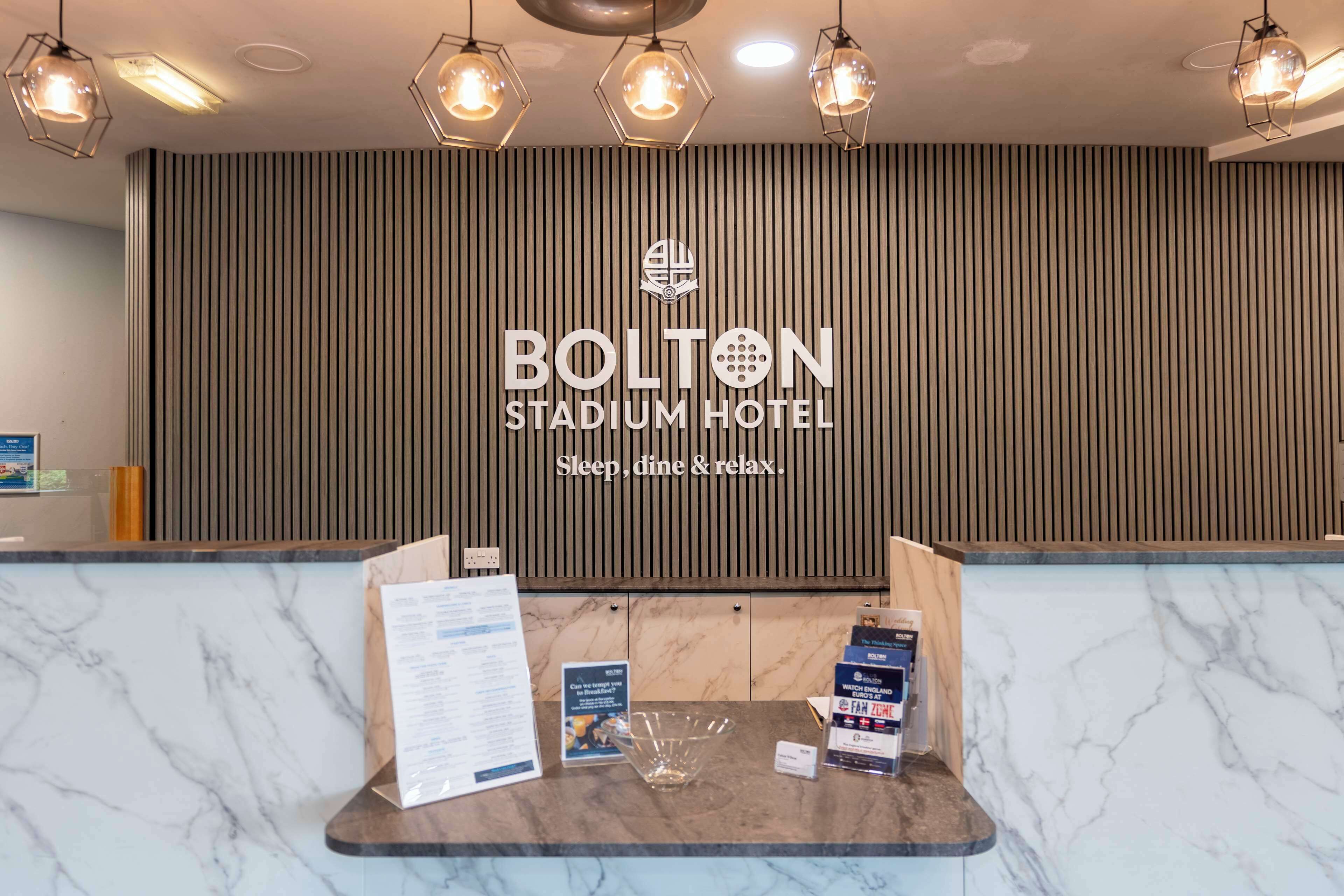 A reception desk at the Bolton Stadium Hotel with a modern design, featuring a marble countertop, hanging pendant lights, and a vertical striped wall behind.