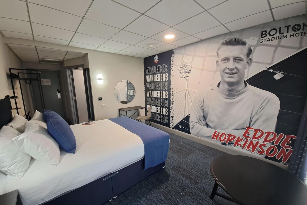 A hotel room with a bed, a desk, a chair, and a large poster of Eddie Hopkinson.