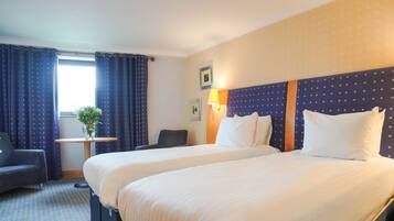 A hotel room with two beds, a small table with a vase, a chair, and a window with curtains.