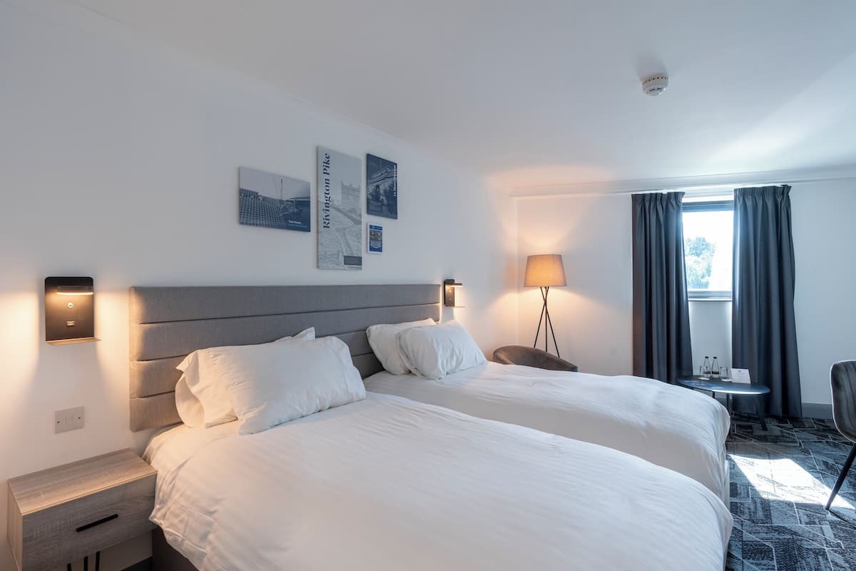 A hotel room with two beds, a bedside table, a lamp, a chair, and a window with curtains.