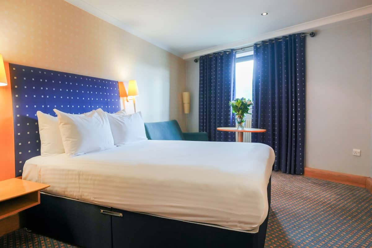 A hotel room with a large bed, two bedside lamps, a small table with a vase of flowers, and a window with curtains.