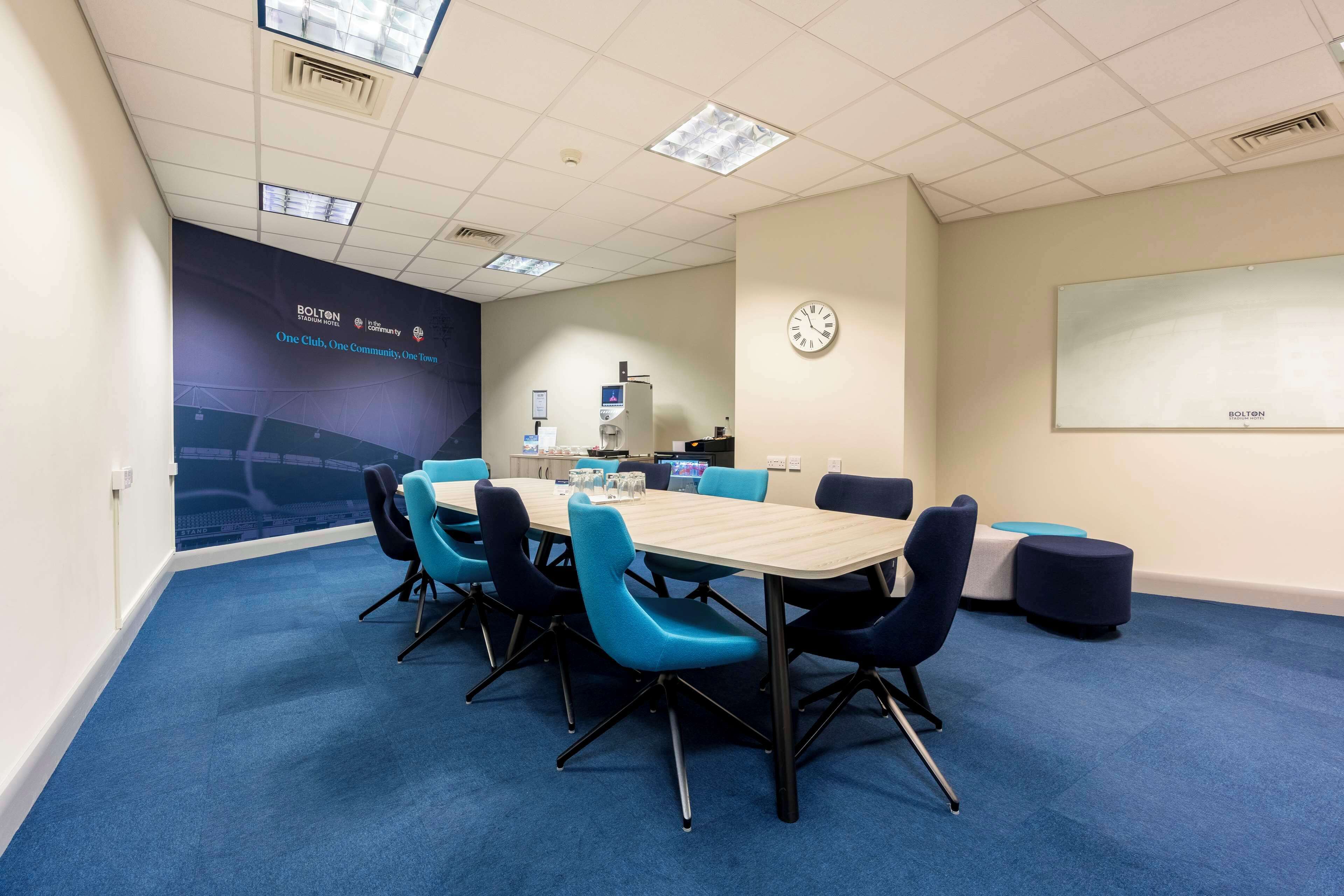 A conference room with a long table, blue chairs, a clock, and a projector screen.