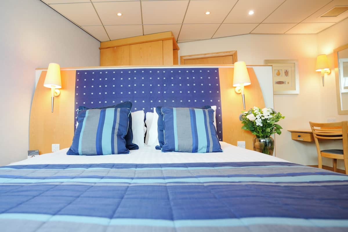 A hotel room with a large bed, two bedside lamps, a wooden headboard, a desk with a chair, and a vase with flowers.