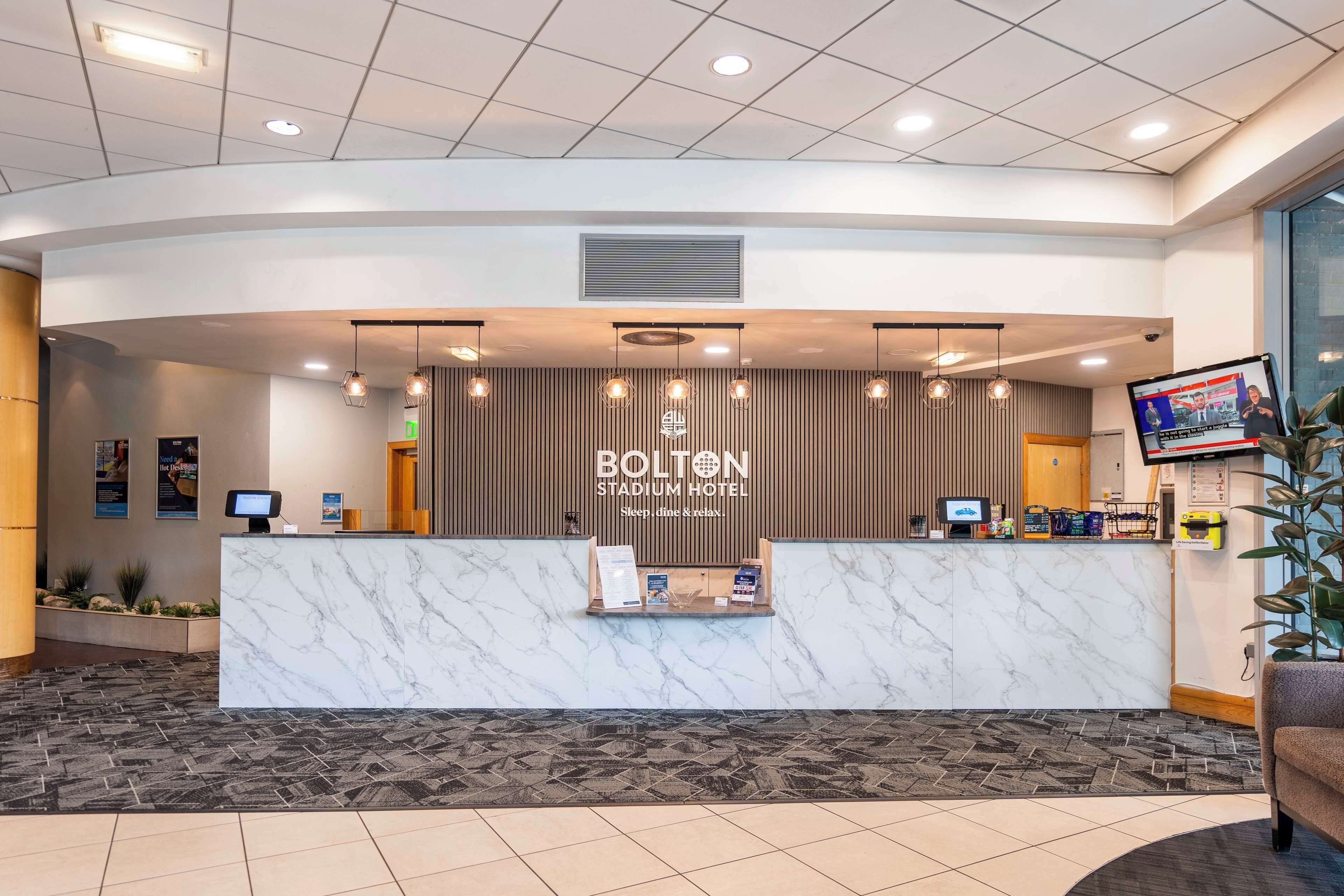 Una zona reception con un bancone in marmo, schermi digitali e un cartello per l'Hotel Bolton Stadium.