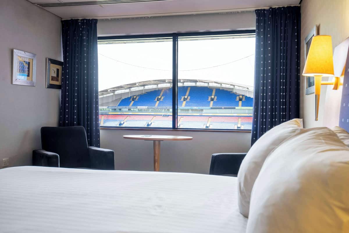 A hotel room with a bed, two chairs, a small round table, and a window with a view of a stadium.