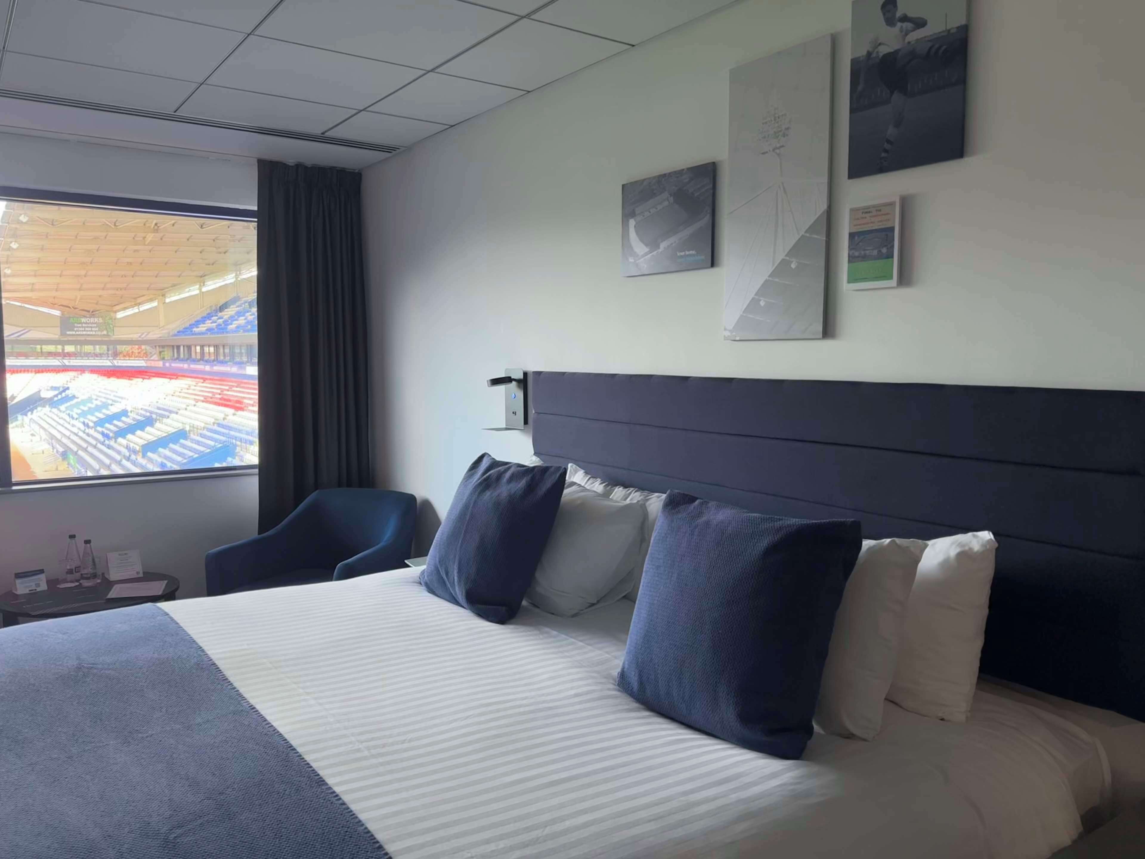 Hotelkamer met een groot bed, uitzicht op een stadion door het raam, een bureau met stoel en een aan de muur bevestigde televisie.