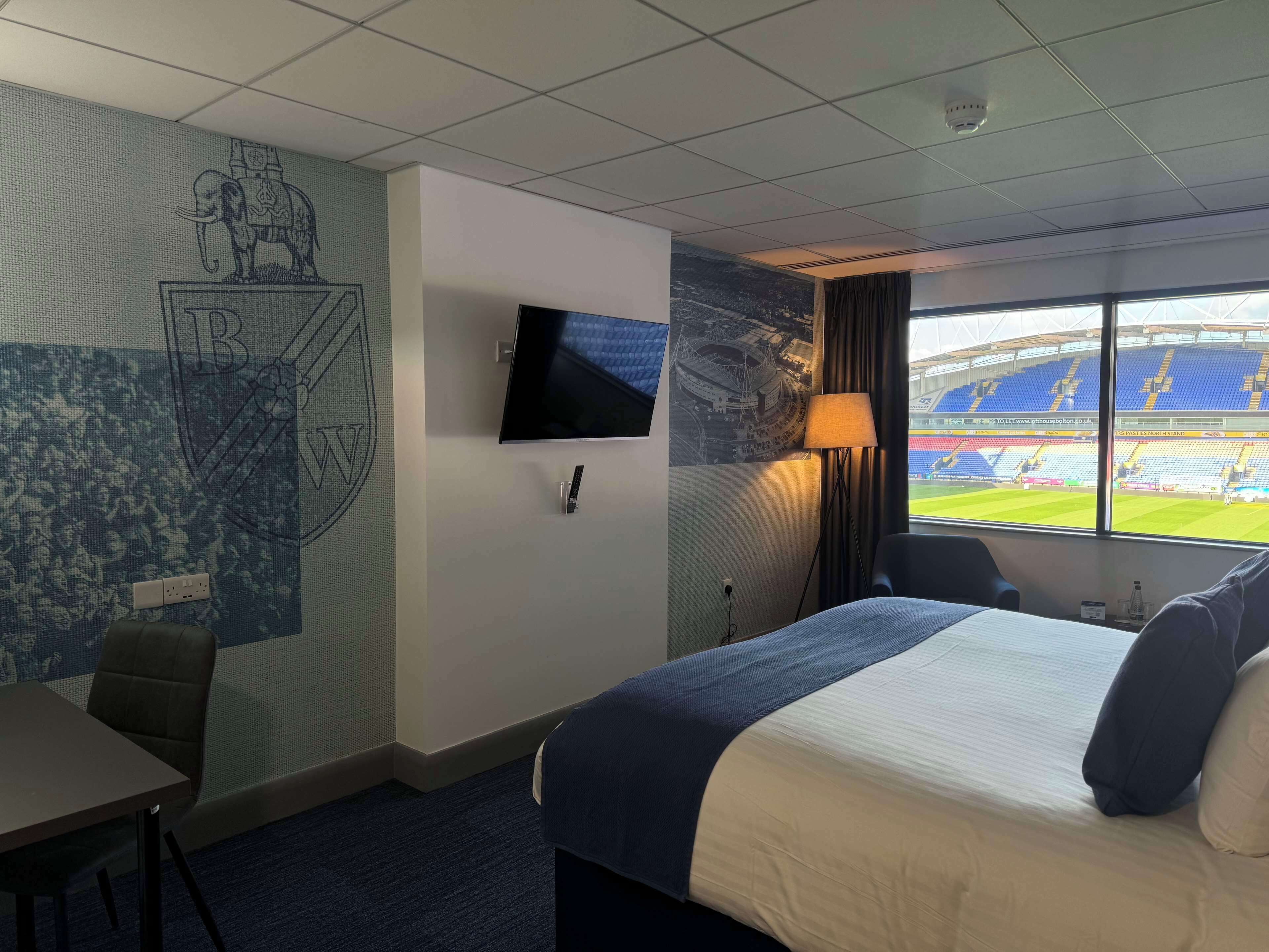 Habitación de hotel con cama, escritorio, silla, televisor y ventana con vista a un estadio.