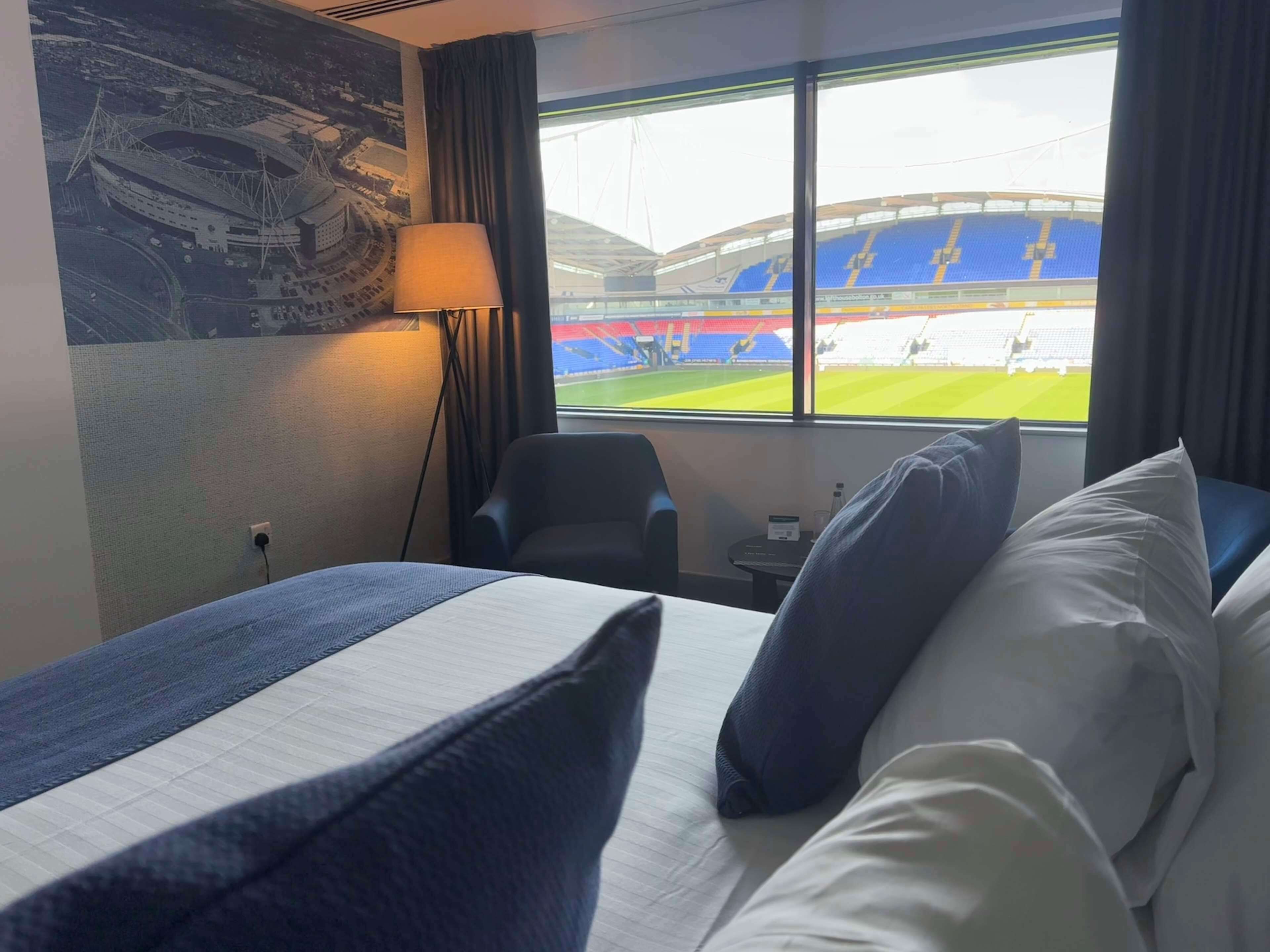 Hotelkamer met een bed, een stoel, een lamp, een klein tafeltje en een raam met uitzicht op een stadion.