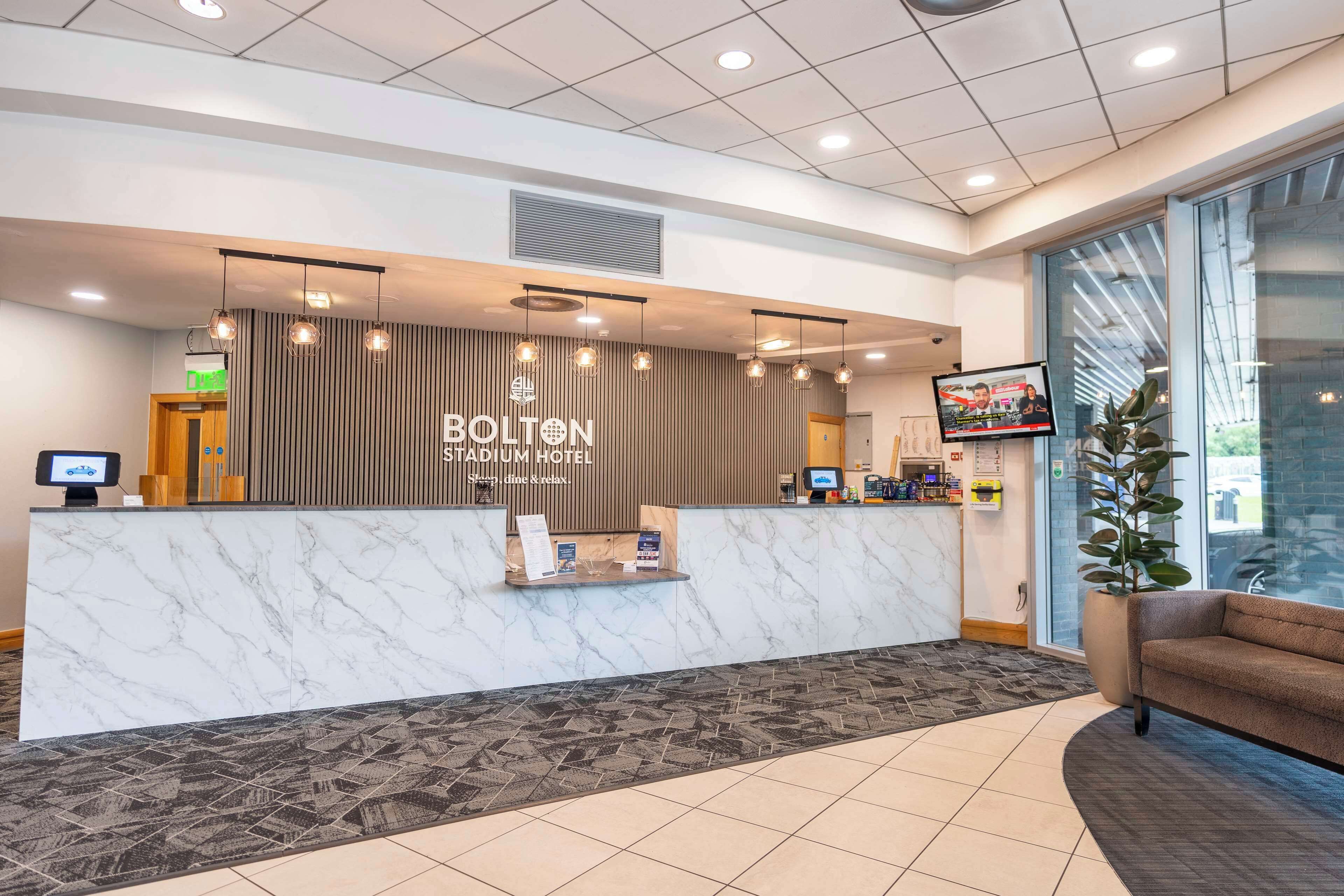 A hotel reception area with a marble counter, a television screen, and a seating area with a plant.
