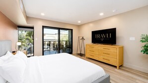 Une chambre moderne avec un grand lit, un téléviseur posé sur une commode en bois et un balcon avec un coin salon extérieur.
