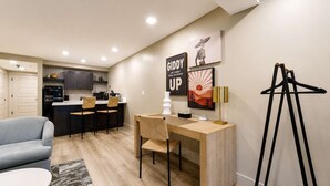 Una cocina moderna con gabinetes oscuros, una zona de comedor con una mesa y sillas de colores claros, y un escritorio pequeño con una lámpara.