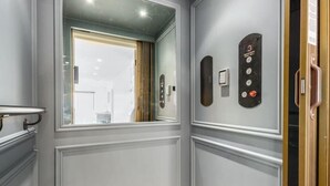 En gang med en dør, der fører til et værelse, et vægmonteret elevatorpanel, og et vindue med udsigt.