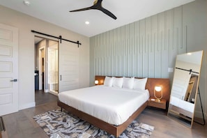 A bedroom with a large bed, a ceiling fan, a bedside table with a lamp, a mirror, and a patterned rug.