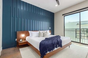A modern bedroom with a large bed, a nightstand with a lamp, and a view of the outside through a large window.