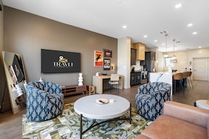 A modern hotel room with a seating area, a dining table, and a kitchen in the background.