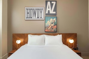 A hotel room with a bed, two bedside lamps, a framed picture of a man in a hat, and two framed signs reading 'HOWDY' and 'AZ'.