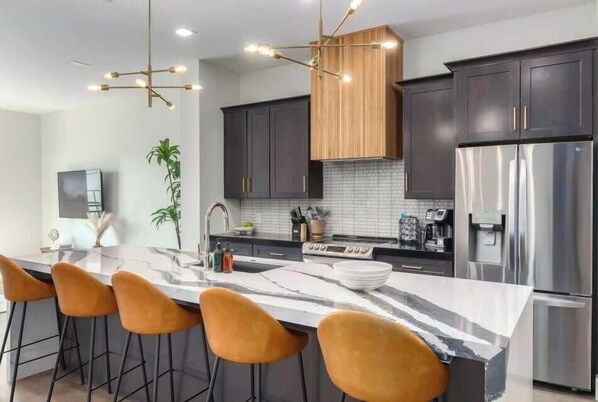 A modern kitchen with dark cabinets, a large island with bar stools, a stainless steel refrigerator, and a pendant light.