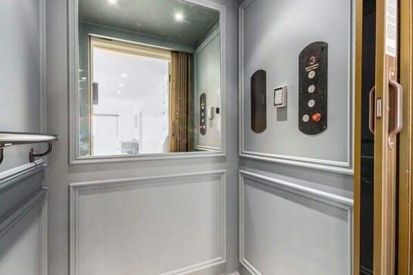 Un pasillo con una puerta que da a una habitación, un panel de control de ascensor en la pared y un ventanal con vistas al exterior.