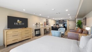 Habitación de hotel moderna con una cama grande, televisión, cómoda de madera, escritorio, silla, kitchenette pequeña y una planta en maceta.