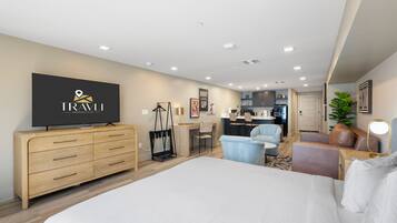 Habitación de hotel moderna con una cama grande, televisión, cómoda de madera, escritorio, silla, kitchenette pequeña y una planta en maceta.