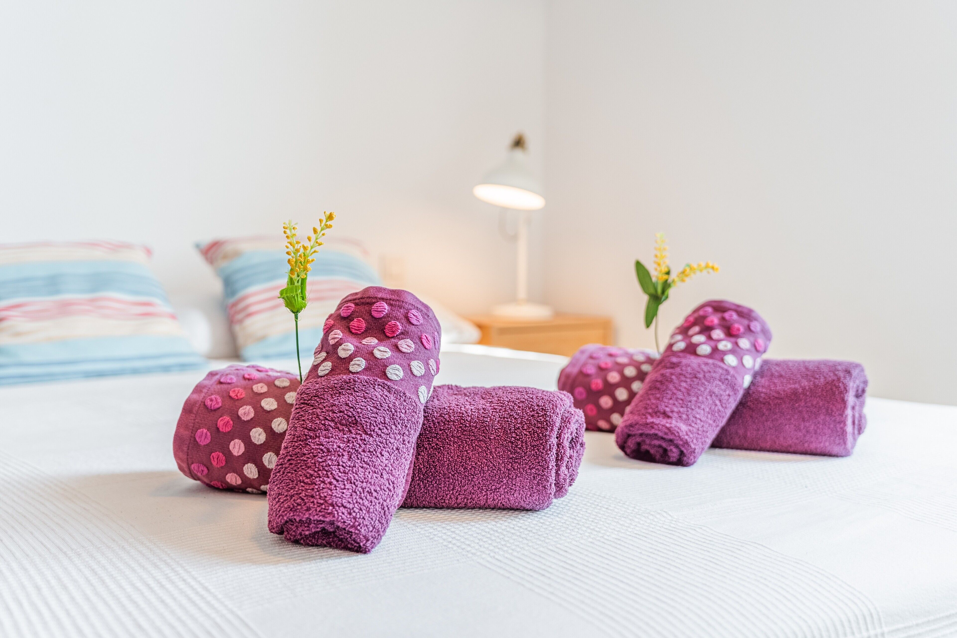 A neatly made bed with rolled towels and decorative pillows, a bedside lamp, and striped pillows in the background.