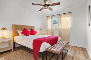 Un dormitorio con cama, mesitas de noche, un banco, ventilador de techo y ventana con cortinas.
