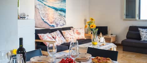 A dining table with plates, glasses, and food, a sofa with floral cushions, and a painting of a beach scene.