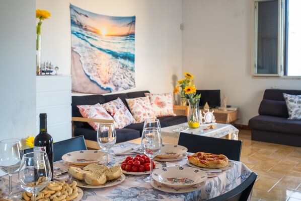 A dining table with plates, glasses, and food, a sofa with floral cushions, and a painting of a beach scene.