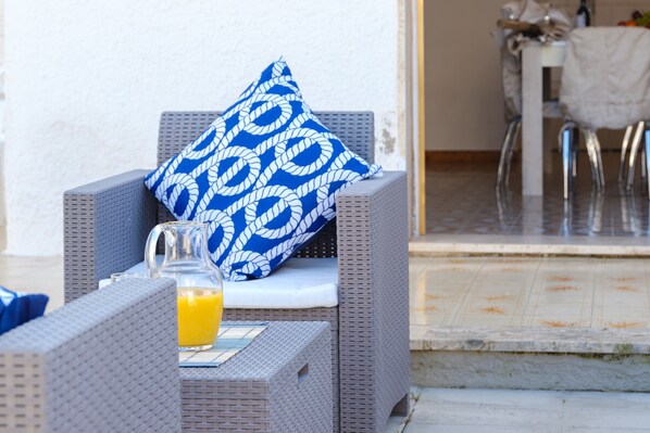 Una silla de mimbre con un cojín blanco y una almohada de estampado azul, una jarra de vidrio con jugo de naranja y una mesa con mantel blanco.
