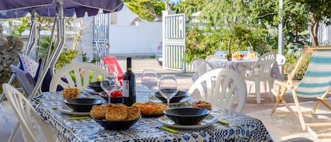An outdoor dining area with a patterned tablecloth, white chairs, and a table set with food and drinks.