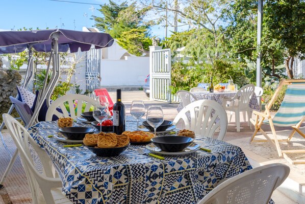 An outdoor dining area with a patterned tablecloth, white chairs, and a table set with food and drinks.