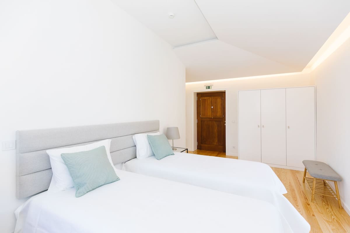 A modern hotel room with two beds, a wooden floor, a wardrobe, and a small stool.
