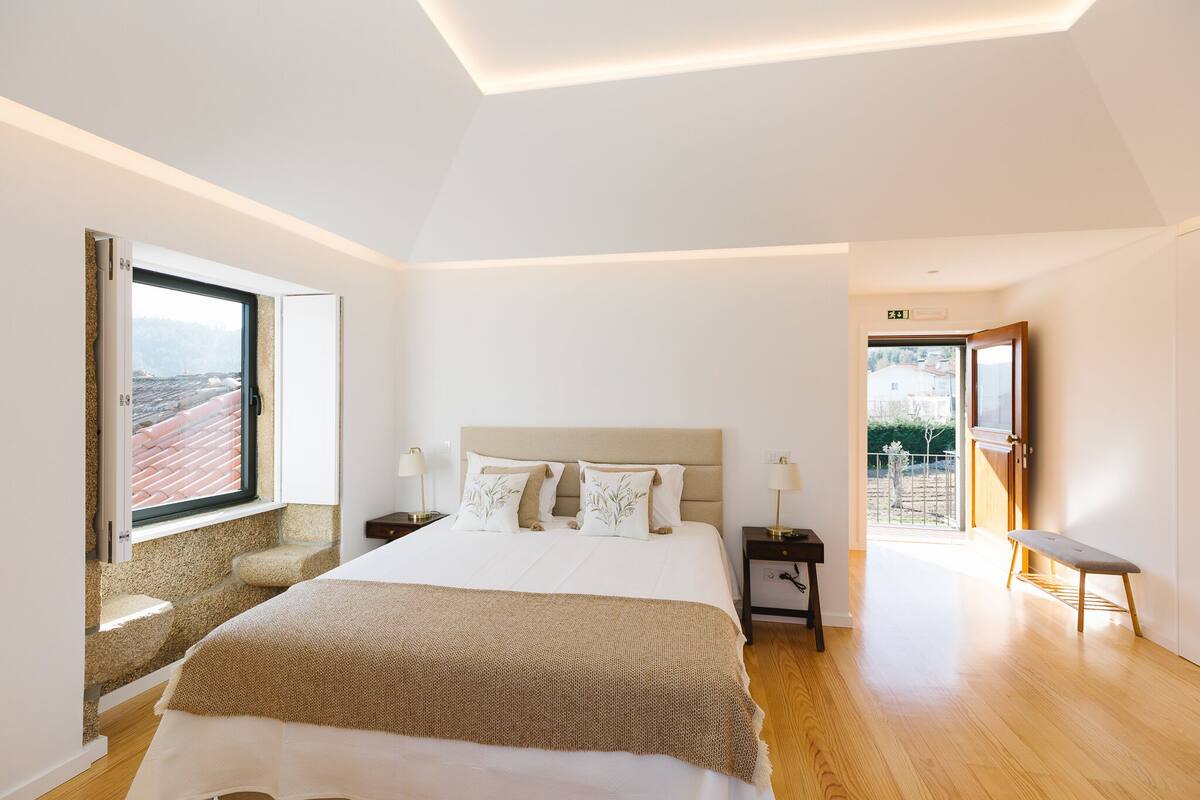 A bedroom with a large bed, a window with a view, a bench, and a door leading to an outdoor area.