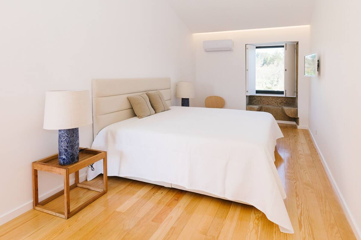 A modern bedroom with a large bed, a bedside table with a lamp, a window with a view, and a built-in bench.