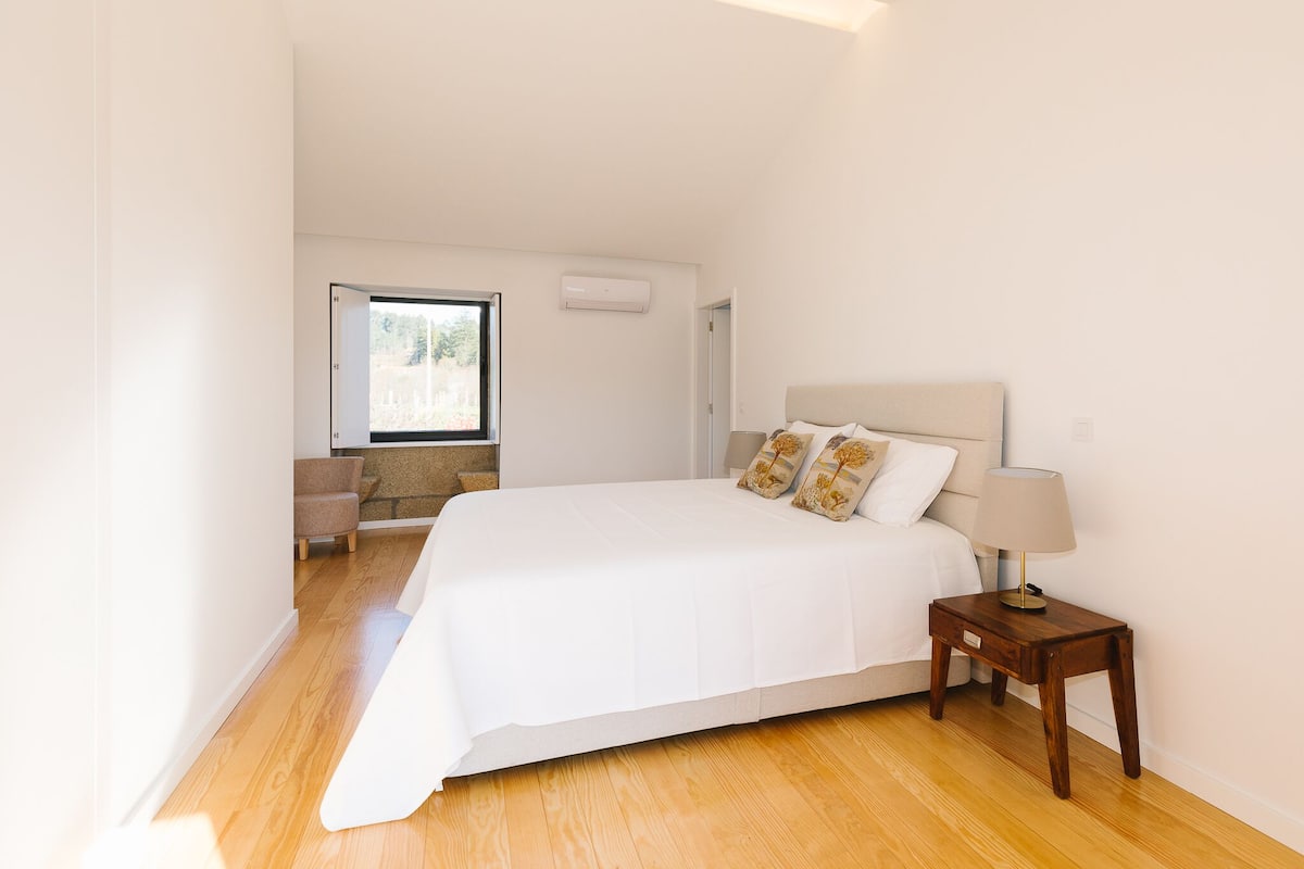 A bedroom with a large bed, a nightstand with a lamp, a window with a view, and a door leading to another room.
