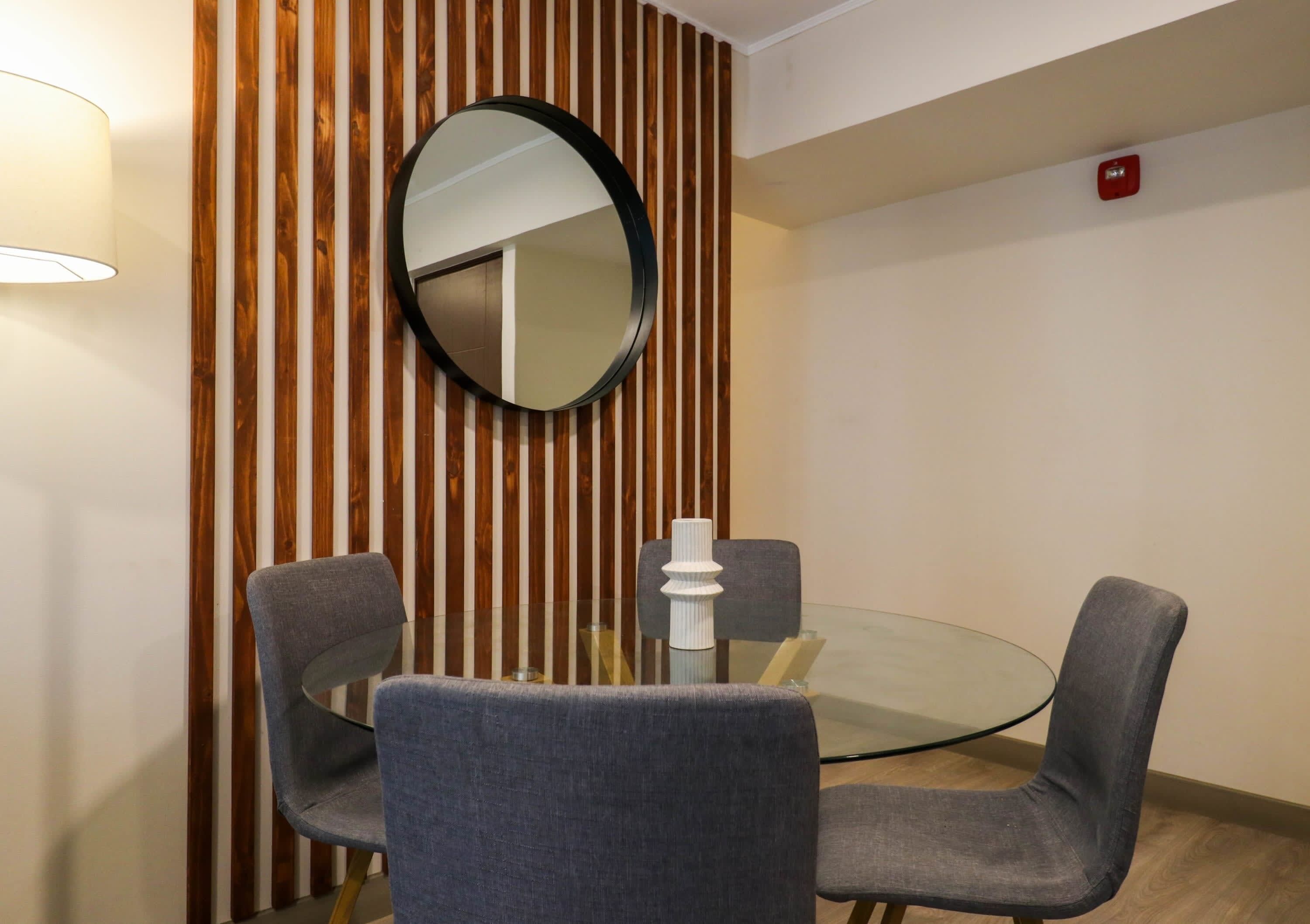 Une salle de conférence avec une table ronde en verre, quatre chaises grises rembourrées, un miroir rond accroché au mur et un claustra en bois.