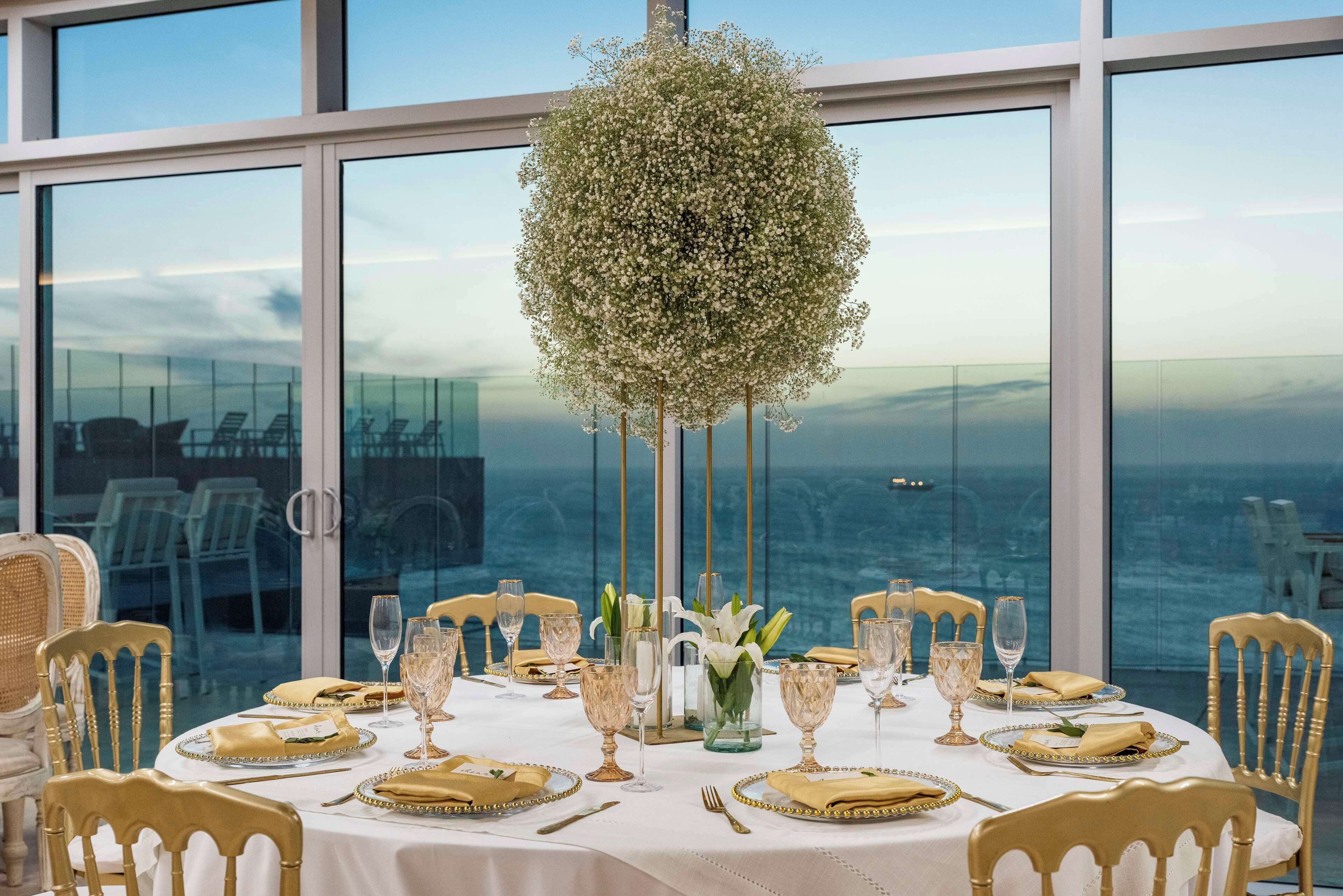 A dining table set with gold and white tableware, surrounded by chairs, with a view of the sea through large windows.
