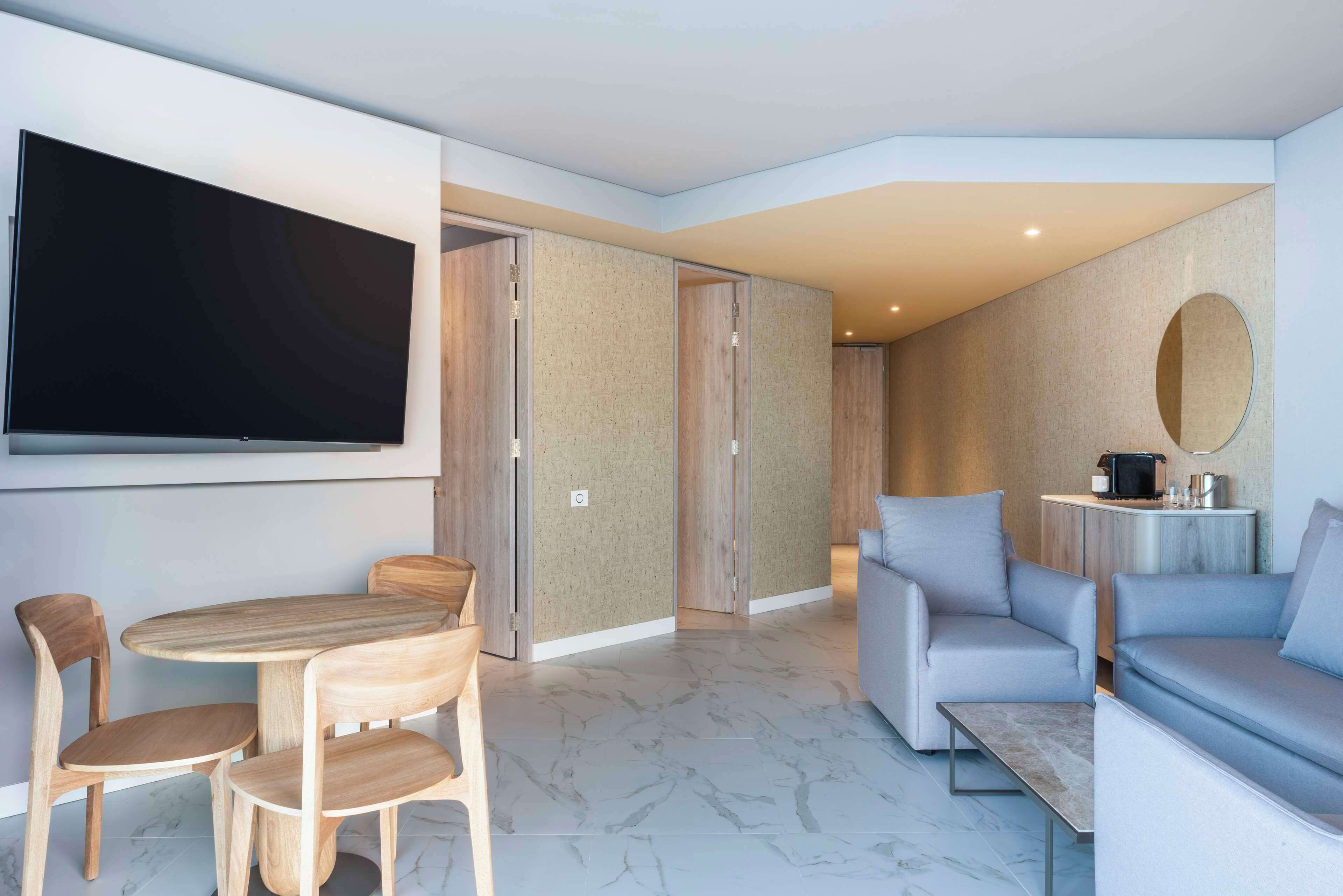 A modern living room with a flat-screen TV, a wooden dining table with chairs, a grey sofa, and a small coffee table.