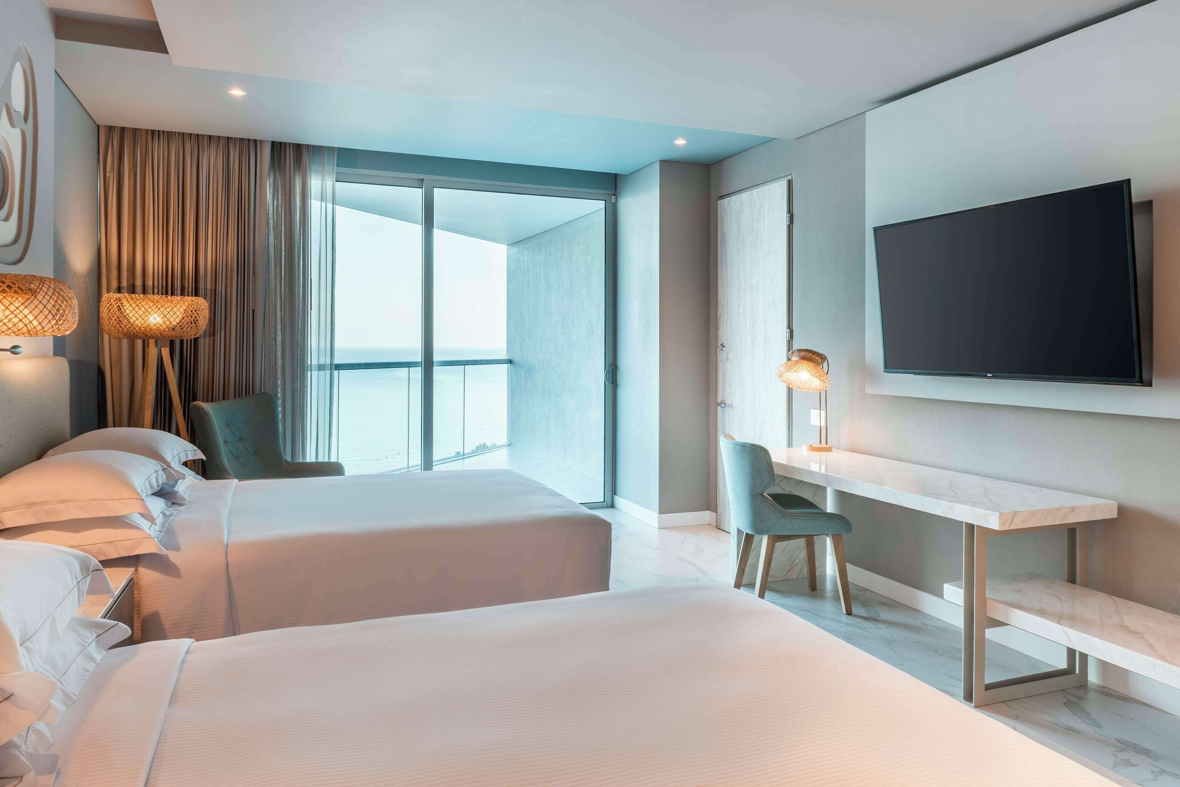 A modern hotel room with two beds, a desk, a TV, and a large window with curtains.