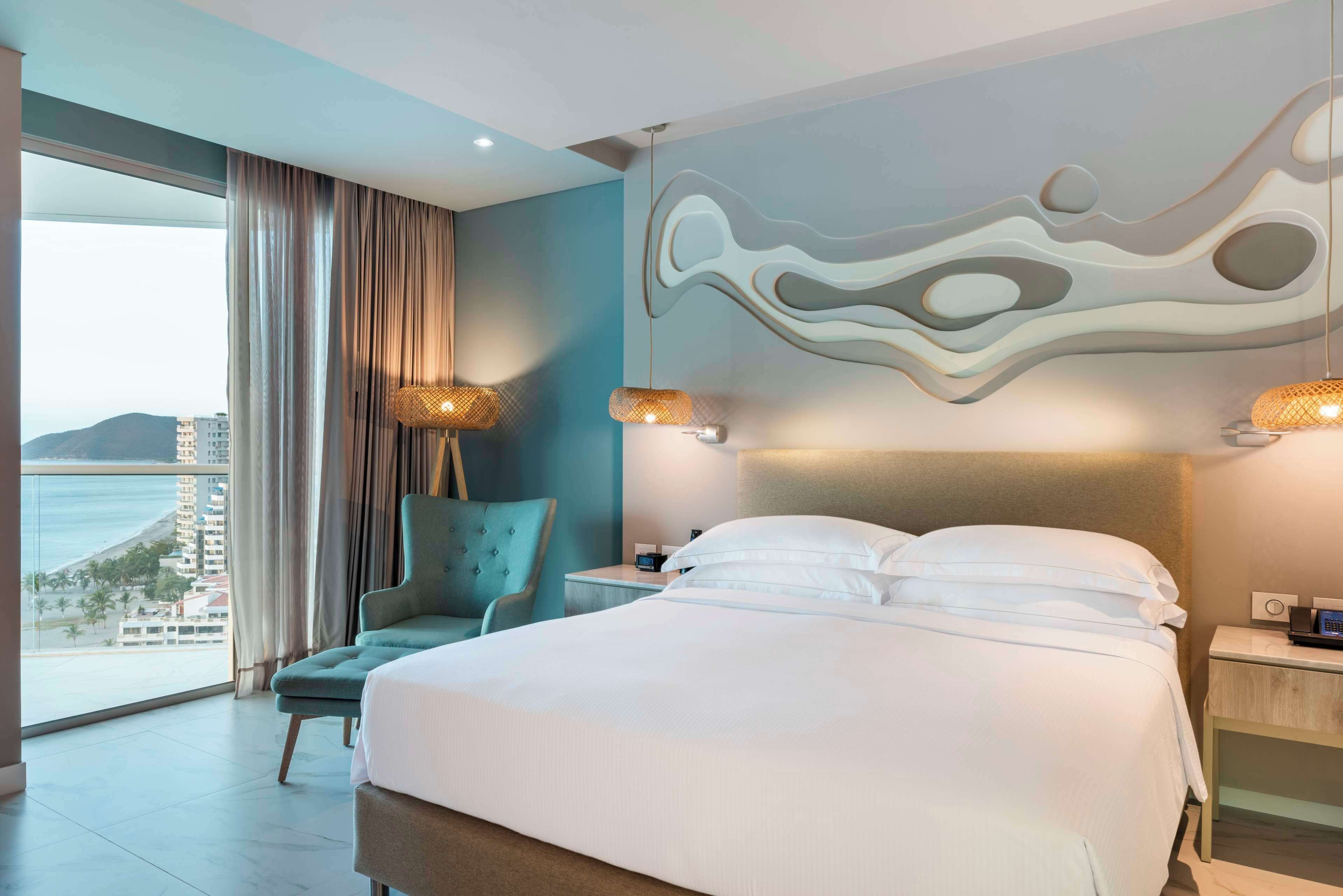 Una habitació d'hotel amb un llit gran, vistes a l'oceà, una cadira i una tauleta de nit amb una làmpada.