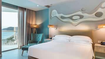 Habitación de hotel con una cama grande, vista al océano, una silla y una mesita de noche con lámpara.