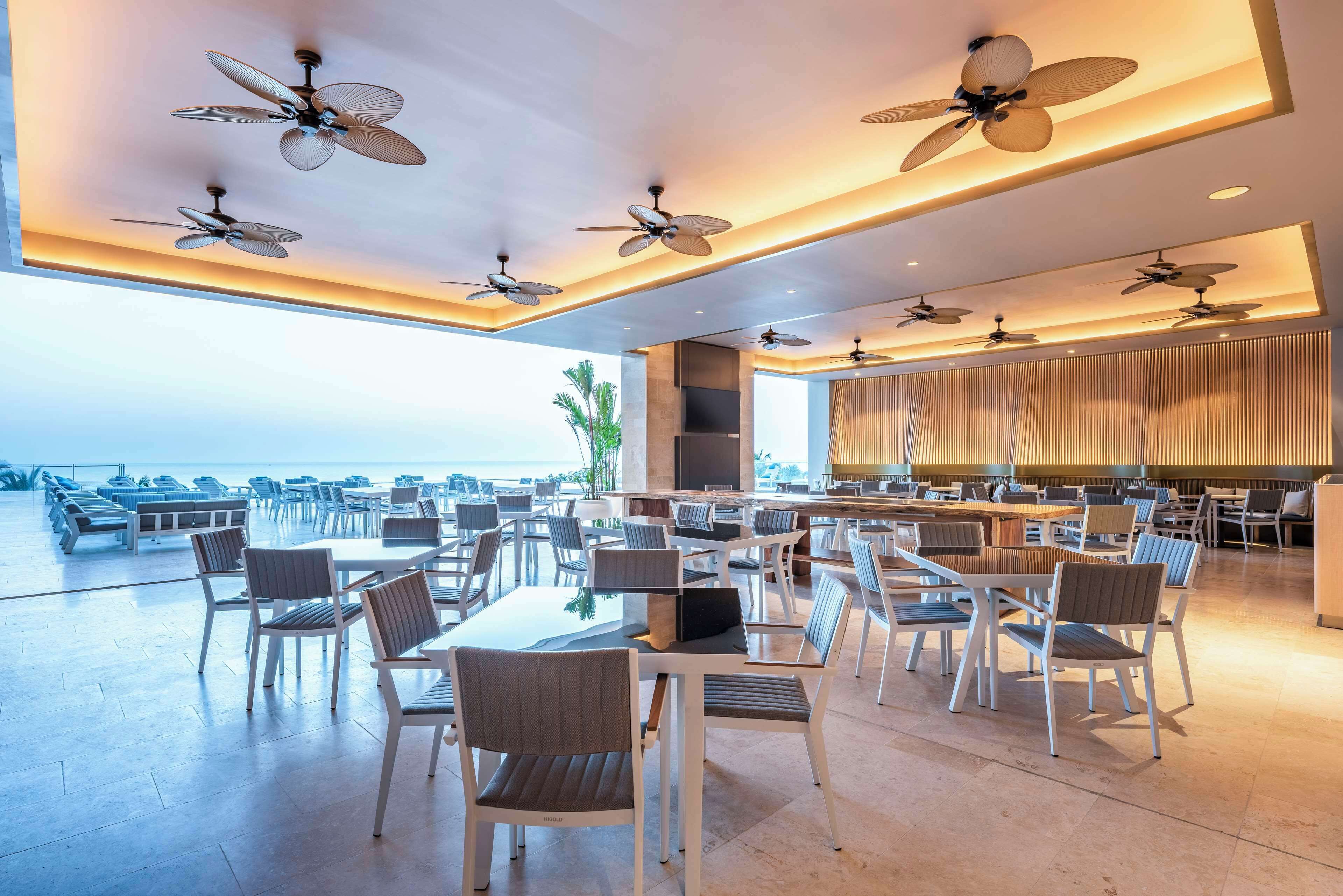 A spacious outdoor dining area with multiple tables and chairs, ceiling fans, and a view of the ocean.