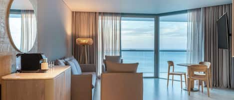 A modern hotel room with a large window, a round mirror, a sofa, a dining table with chairs, and a flat-screen TV.